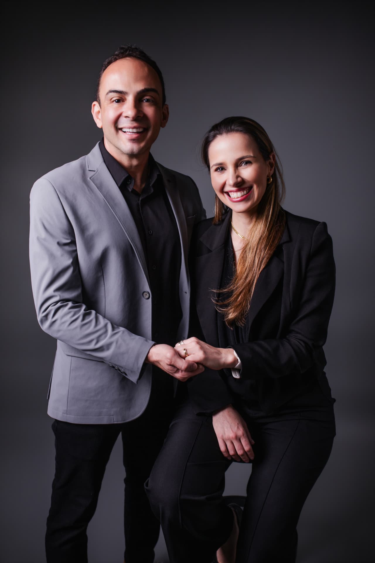 Dr. Francis Pelegrini e Dra. Andressa Rossetti Pelegrini sorrindo lado a lado, de mãos dadas, em fundo cinza escuro