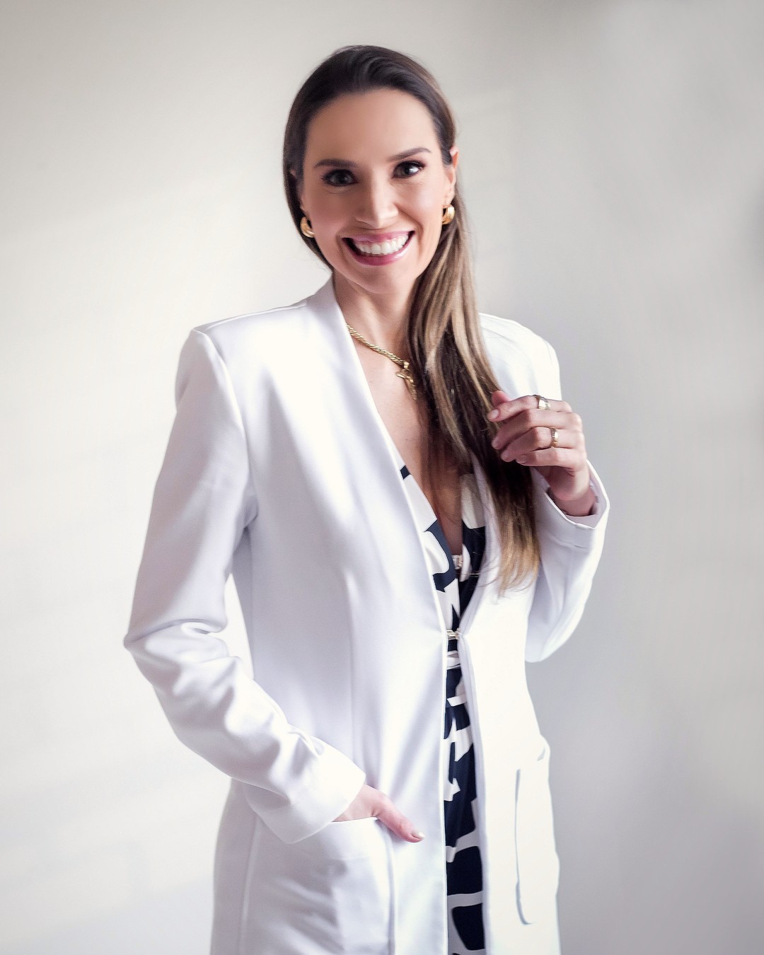 Dra. Andressa Rossetti Pelegrini sorrindo, de jaleco branco, em pé com uma mão no bolso do jaleco, em fundo claro neutro