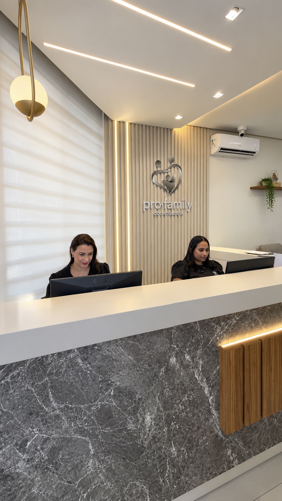 Recepção da Pro Family Odontologia com balcão de mármore, painel ripado iluminado e duas recepcionistas atendendo